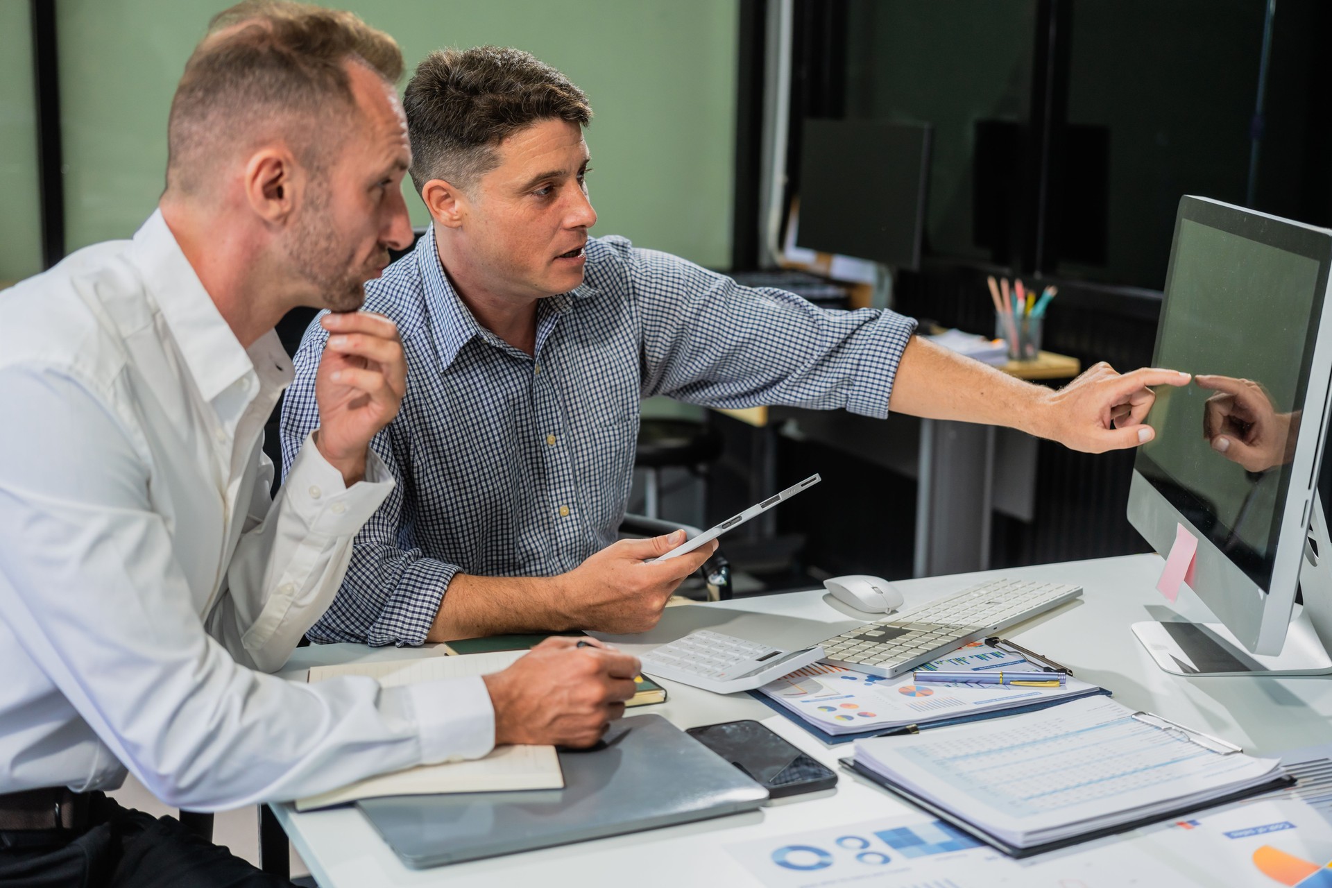 Caucasian middle-aged male businessperson and an Italian accountant are seated at a desk, engaging in a professional meeting, discussing various aspects of technology and innovation in their fields.